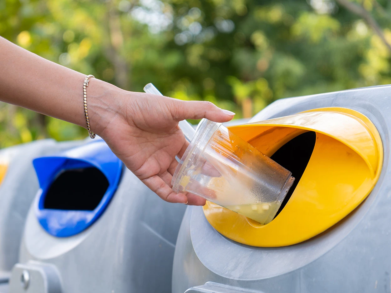 Eine Hand wirft einen Plastikbecher in einen Mülleimer mit einer gelben Öffnung, währen links daneben ein Mülleimer mit einer blauen Öffnung steht.